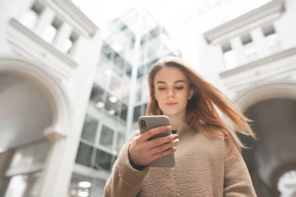 Κομψό κορίτσι απολαμβάνει ένα smartphone στο φόντο μιας σύγχρονης αστικής αρχιτεκτονικής. Νεαρή επιχείρηση γυναίκα με ένα smartphone στα χέρια της στο φόντο ενός σύγχρονου κτιρίου. Εστίαση στο smartphone — Φωτογραφία Αρχείου