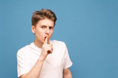 Beyaz bir tişört adam dudaklarına parmağını koydu ve mavi bir arka planda izole, sessizlik bir işareti gösterir. Adam diyor ki: sessiz olun lütfen. Ciddi genç adam, bir jest konuşmayın sorar