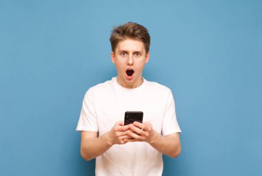 Elinde bir akıllı telefon ile mavi bir arka planda duran ve açık bir ağız ile kameraya bakarak şok bir genç adamın portresi. Elinde bir smartphone ile hayran çocuk, izole