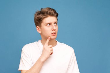 Beyaz bir tişört bir adam portre mavi bir arka planda duruyor, bir kenara bakar ve düşünüyor. Düşünceli genç adam mavi bir arka planda pozlar ve reklam için bir yere bakar. Izole.