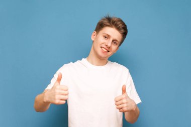 Beyaz bir tişört mutlu genç adam parmaklarını gösterir, gülümsüyor ve mavi bir arka planda kamera bakıyor. Pozitif genç adam gibi bir jest gösterir. Izole