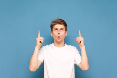 Beyaz bir T-shirt içinde sürpriz genç adam parmaklarını gösterir ve reklam için boş bir alana bakıyor, mavi bir arka planda izole. Boşaltmak