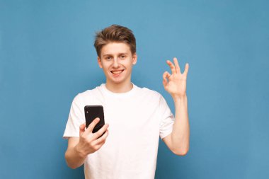 Mutlu genç adam mavi bir arka planda duruyor, elinde bir akıllı telefon tutan, bir işaret ok gösterir, kamera içine bakar ve gülümsüyor. Elinde telefon ile pozitif adam mavi arka planda izole edilir.