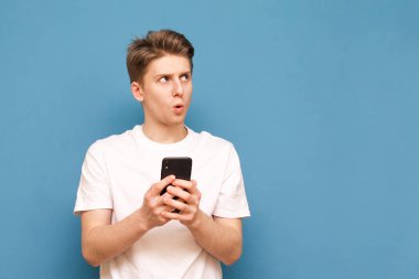 Beyaz bir tişört sürpriz çocuk elinde bir smartphone tutar ve mavi bir arka planda bir kenara bakar. Duygusal genç adam bir akıllı telefon kullanır ve bir kenara görünüyor, izole. Boşaltmak