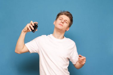 Young man uses perfumes on a blue background wearing a white T-shirt. Spray perfume from the bottle and enjoy the smell with closed eyes. clipart