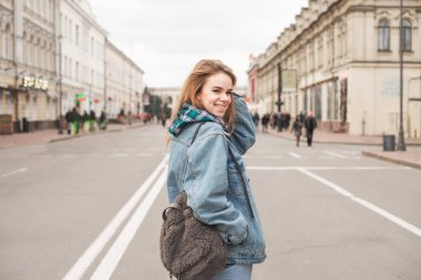 Casual giyim güzel Bayan kamera bakarak ve gülümseyen bir sokak peyzaj arka planda bir sırt çantası ile ayakta. Güzel bir şık kız sokak portresi. Sokak modası