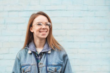 Mavi bir arka planda mutlu bir kızın yakın çekim portre, yan ve gülümseyen, bir kot ceket ve gözlük giyiyor. Rahat kıyafetler hipster kız mavi bir duvarın arka planda pozlar. Boşaltmak