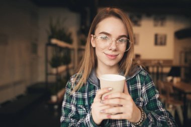 Rahat bir kahve dükkanında elinde bir fincan kahve ile sevimli kız, bir gömlek ve Casual giyim giyen, kamera bakıyor ve gülümsüyor. Yakın yukarı portre