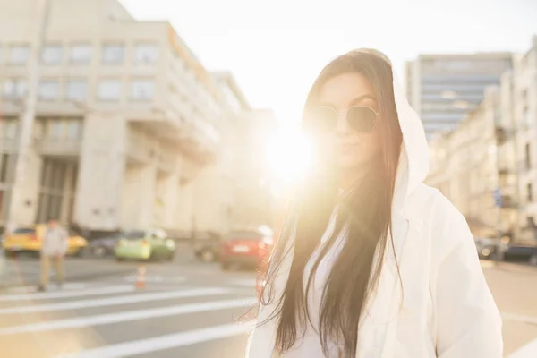 Ragazza alla moda in un abito casual bianco sullo sfondo del tramonto, paesaggio stradale. Elegante donna in occhiali da sole ritratto con retroilluminazione sulla strada di megapoli . — Foto Stock