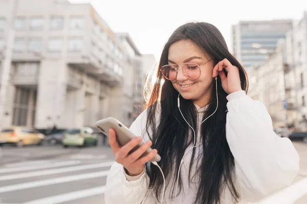 Улыбающаяся девушка в белой повседневной одежде и солнцезащитных очках, слушает музыку в наушниках, смотрит на смартфон в руке и улыбается. — стоковое фото