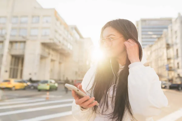 Μοντέρνα γυναίκα στέκεται στο παρασκήνιο μιας πόλης, smartphone στο χέρι της, φοράει ένα λευκό μπουφάν και ακούει τη μουσική. Κορίτσι του δρόμου πορτραίτο στα ακουστικά στο φόντο της μεγαλολογία — Φωτογραφία Αρχείου