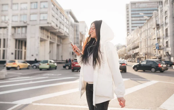 Attractive brunette woman spectacles searching songs in network, listening to music in the headphones. Stylish girl in a white jacket walking along the street with a smartphone — Stock Photo, Image