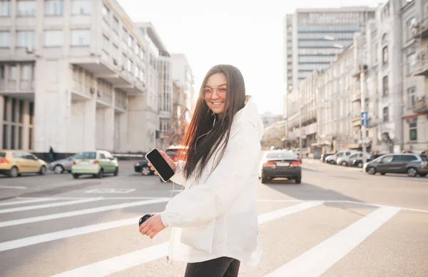 Menina feliz em roupas casuais e óculos de sol com um smartphone na mão, desce a rua contra a cidade durante o sol, olha câmera e sorri. Retrato de uma morena na moda em um passeio — Fotografia de Stock