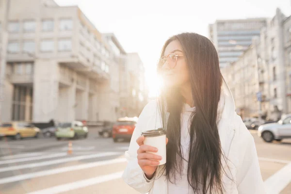 Πορτρέτο του δρόμου από ένα κομψό μελαχρινή κορίτσι, φορώντας ένα λευκό μπουφάν, κρατά ένα φλιτζάνι καφέ στα χέρια της, στέκεται στο φόντο της πόλης το ηλιοβασίλεμα. Μοντέρνα γυναίκα που περπατάει στο δρόμο — Φωτογραφία Αρχείου