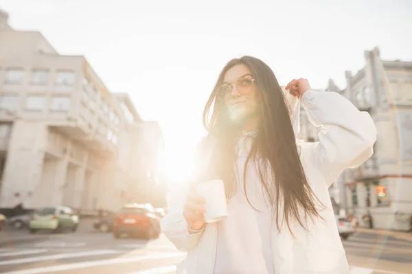 Φωτογραφία του δρόμου από μια κομψή γυναίκα με ένα λευκό μπουφάν, κρατώντας ένα φλιτζάνι καφέ στα χέρια του και ποζάρει στο φόντο του τοπίου της πόλης στο ηλιοβασίλεμα, κοιτάζοντας πλάγια και διορθώνοντας το καπό — Φωτογραφία Αρχείου
