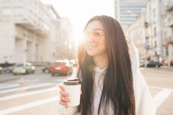 Ευτυχισμένο κορίτσι με ένα φλιτζάνι καφέ στα χέρια της στέκεται στο δρόμο στο φόντο του ηλιοβασιλέματος, κοιτάζει πλάγια και χαμόγελα, φοράει ένα κομψό λευκό μπουφάν και γυαλιά ηλίου. Χαμογελαστή μελαχρινή, πορτρέτο του δρόμου — Φωτογραφία Αρχείου