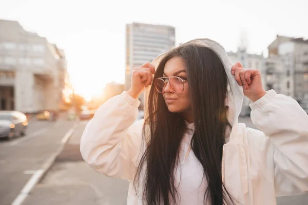 Portret van een brunette meisje in de zonnebrillen en een wit jasje staande tegen de achtergrond van de zonsondergang en op zoek zijwaarts. Modieus meisje in een casual kleding straat portret — Stockfoto