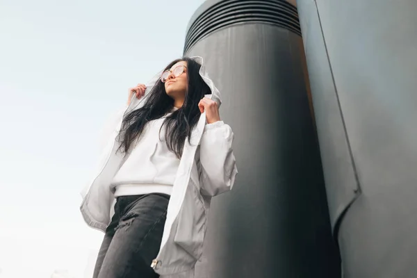 Ritratto alla moda di una ragazza in un abbigliamento casual bianco e occhiali, pose sulla fotocamera vicino alle colonne nere, vista dal basso. Foto urbana di una ragazza alla moda . — Foto Stock