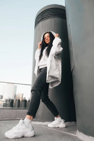 Retrato callejero de moda de una chica vestida con ropa casual, con una chaqueta blanca, zapatillas y gafas, de pie a toda altura, posa y mira a la cámara. Moda callejera. Foto vertical . — Foto de Stock