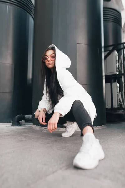 Street fashion portrait model in a stylish casual dress and pink glasses poses a futuristic urban location. Stylish girl sat down on the ground, looking at the camera , modern architecture — Stock Photo, Image