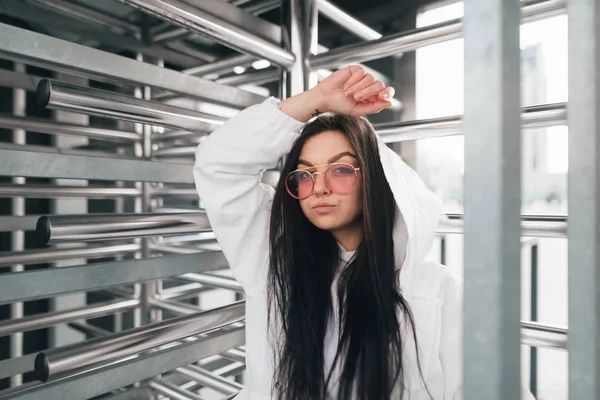 Retrato de rua de uma menina atraente em roupas de rua perto de catracas. Modelo de menina elegante em óculos rosa e uma jaqueta branca posa para uma localização futurista. Conceito de moda rua . — Fotografia de Stock