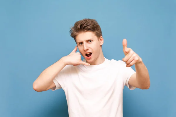 Ring mig tillbaka snälla. Ung man med en "Call Me"-gest visar ett finger och tittar på kameran på en blå bakgrund. Guy och en vit T-shirt vill kalla honom, visar en praktisk gest av samtalet — Stockfoto