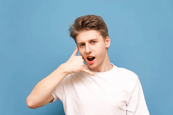 Positiv ung mand i en hvid T-shirt ser på kameraet og viser en hands-on gestus af opkald, isoleret på en blå baggrund. Ring til mig. Copyspace - Stock-foto