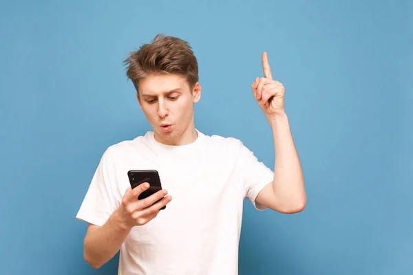 El tipo con una camiseta blanca sostiene un teléfono inteligente en sus manos, mira la pantalla con ojos asombrados y muestra su dedo hacia un espacio en blanco, aislado sobre un fondo azul. Espacio de copia — Foto de Stock