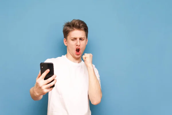 Portrait of a joyful man with a smartphone in his hands, emotionally reacts, looks at the screen, wears a white T-shirt and stands on a blue background. Copyspace — Stock Photo, Image