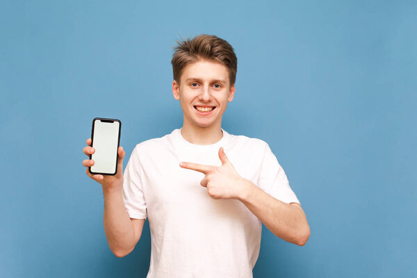 Happy young man in a white T-shirt holds a smartphone in his hands, shows his finger on the white screen, looks into the camera and smiles on a blue background. Isolated