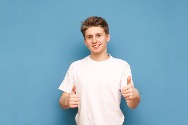 Joven sonriente mostrando los pulgares hacia arriba, mostrando un signo como llevar una camisa blanca y mirando a la cámara sobre un fondo azul. Un hombre feliz con una camiseta blanca muestra los dedos hacia arriba. Espacio de copia —  Fotos de Stock