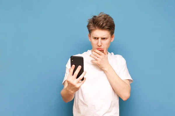 Tipo confundido de pie sobre un fondo azul con teléfono inteligente en la mano y miradas desconcertadas en la pantalla. Chico gracioso en una camiseta blanca utiliza un teléfono inteligente sobre un fondo azul . — Foto de Stock