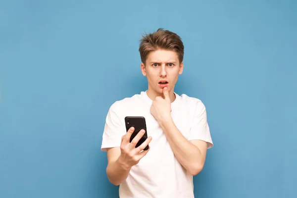 Portrait of a surprised young man standing with a smartphone in his hands on a blue background, looking into the camera. Puzzled guy in a white T-shirt with a smartphone in his hand, isolated — Stock Photo, Image