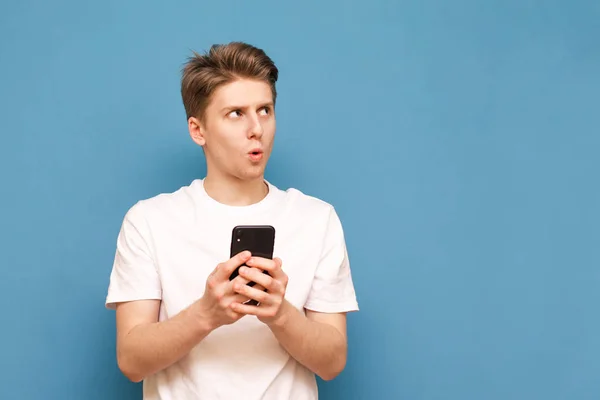 Surprised boy in a white T-shirt holds a smartphone in his hands and looks aside on a blue background. Emotional young man uses a smartphone and looks aside, isolated. Copyspace — Stock Photo, Image