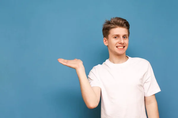 Un joven divertido con una camiseta blanca se levanta sobre un fondo azul y muestra su mano en su lugar para el texto, mira a la cámara y sonríe. Aislado . — Foto de Stock