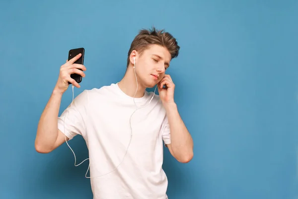 Guapo joven escucha música en sus auriculares con los ojos cerrados, sosteniendo un teléfono inteligente en sus manos y disfrutando de un fondo azul.Guy escucha su música favorita en los auriculares. —  Fotos de Stock