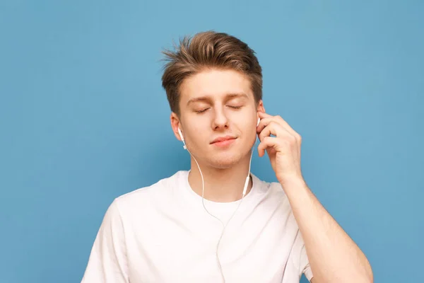 Närbild porträtt av en kille lyssnar på musik i hans hörlurar med ögonen stängda och har en blå bakgrund. Eleven lyssnar på musik i hörlurarna. Isolerade. — Stockfoto
