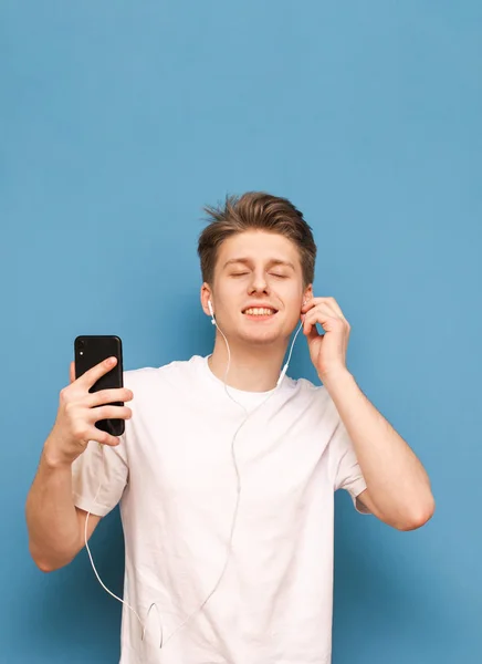 Красивый молодой человек слушает музыку в наушниках с закрытыми глазами, держа в руках смартфон и наслаждаясь синим фоном. Парень слушает свою любимую музыку в наушниках. — стоковое фото
