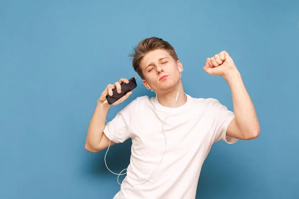 Веселый молодой человек слушает музыку в наушниках и танцует со смартфоном в руке на синем фоне. Парень в белой футболке любит танцевать и слушать музыку в наушниках. Copyspace — стоковое фото