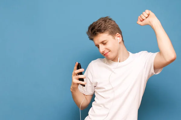 Позитивный парень в белой футболке, танцующий с наушниками в ушах и смартфоном в руке на синем фоне. Молодой человек в наушниках изолирован на синем фоне. Copyspace — стоковое фото