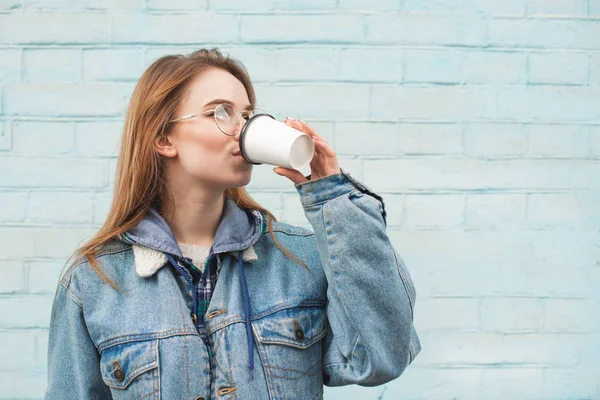 Retrato de uma menina elegante contra o fundo de uma parede azul, olhando para os lados e beber café de uma xícara de papel.Menina bonita em óculos e roupas casuais bebe um copo na rua . — Fotografia de Stock