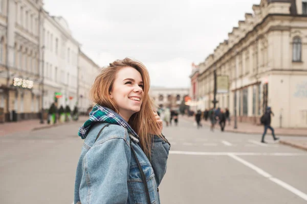 Sorrindo menina atraente andando ao longo da rua da cidade, vestindo um casaco de jeans vestido casual, olhando para os lados, fundo da paisagem da cidade. Estudante bonita nas ruas da cidade — Fotografia de Stock