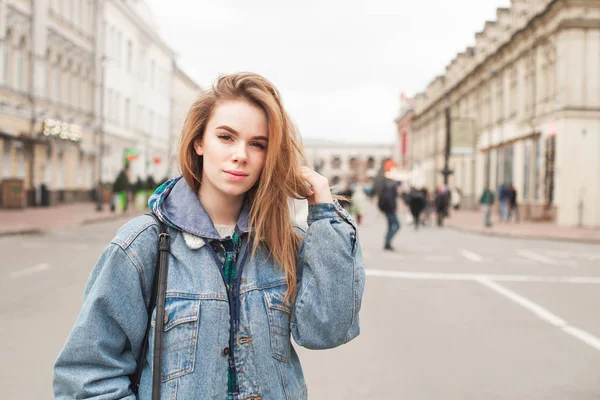 Retrato de rua de uma menina loira atraente vestindo uma jaqueta de ganga, de pé na rua e olhando para a câmera. Menina bonita estudante andando na rua . — Fotografia de Stock