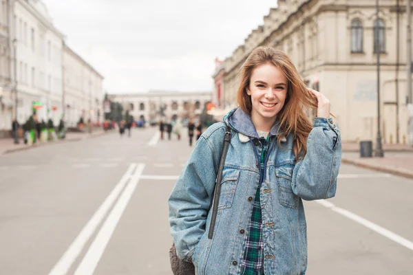 Fille heureuse en vêtements décontractés est debout dans la rue de la ville, souriant et regardant la caméra. Étudiante positive avec un sac à dos contre un paysage de rue. Portrait de rue . — Photo