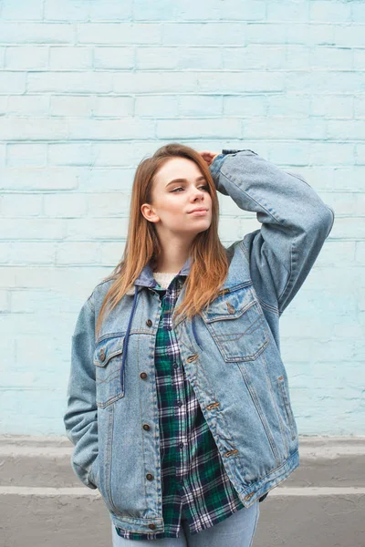 Menina atraente em roupas casuais, vestindo uma jaqueta de ganga, posando contra o fundo de uma parede azul e olhando para longe. Foto vertical — Fotografia de Stock