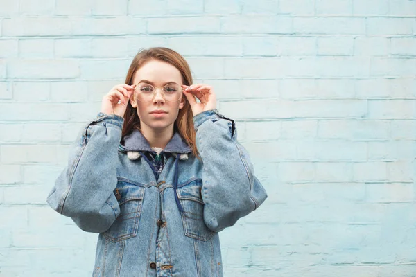 Menina bonita em uma jaqueta de ganga fica contra o fundo de uma parede azul, olha para a câmera e corrige os óculos. Menina elegante hipster posando em um fundo azul. Espaço de cópia — Fotografia de Stock