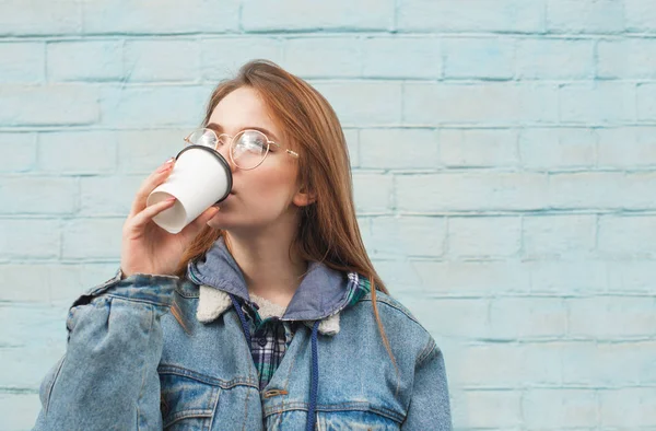 Beautiful girl drinking coffee from a paper cup on the street, wearing a denim jacket, against the background of a blue wall.Hipster Girl in casual clothing drinks coffee on a blue background — стоковое фото