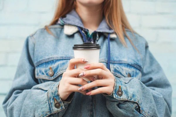Крупным планом девушка в джинсовой куртке держит бумажную чашку с кофе на фоне синей стены. Fantom фото. Copyspace — стоковое фото
