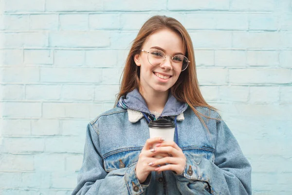 Glimlachend meisje poseert aan de camera op de achtergrond van een blauwe muur, houdt een papieren glas koffie in haar handen, kijkt in de camera en lacht, draagt een jeans jas en een bril. Copyspace — Stockfoto