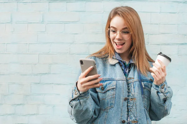 Menina alegre de pé sobre um fundo azul com uma xícara de café e um smartphone na mão, parece surpreendido.Hipster menina vestindo uma jaqueta de jeans usa um smartphone e se alegra, fundo de uma parede azul — Fotografia de Stock